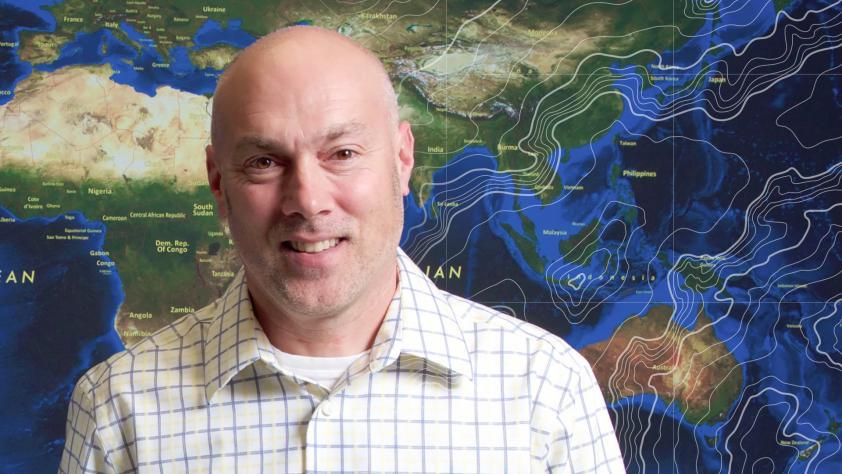 绿帽社 professor of geography Dean Butzow in front of a world map.