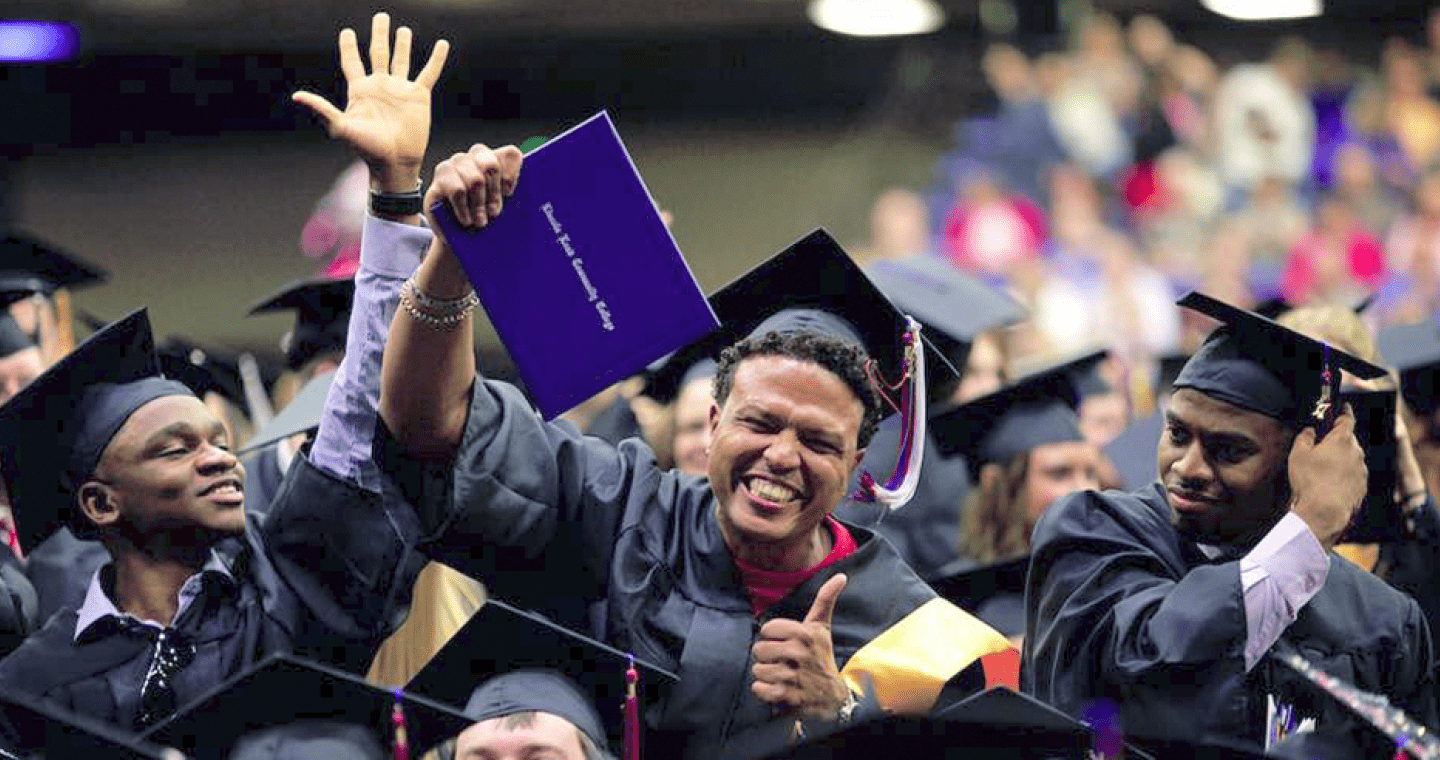 A student wearing a graduation cap and gown holds up a diploma cover and smiles, giving a thumbs up while in a crowd of other graduates.